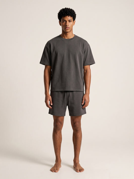 A man modeling a dark gray short-sleeve t-shirt and matching dark gray shorts against a plain, light beige studio background.
