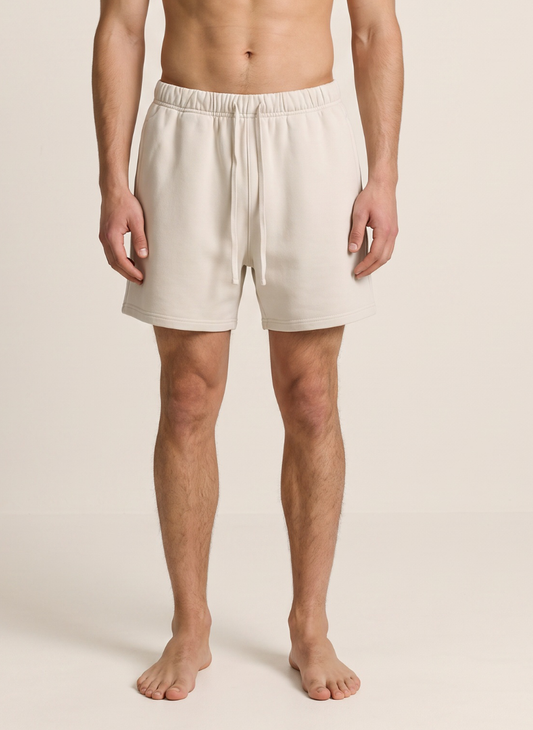 A man wearing light beige drawstring shorts, shown from the chest down against a plain, warm off-white studio background.