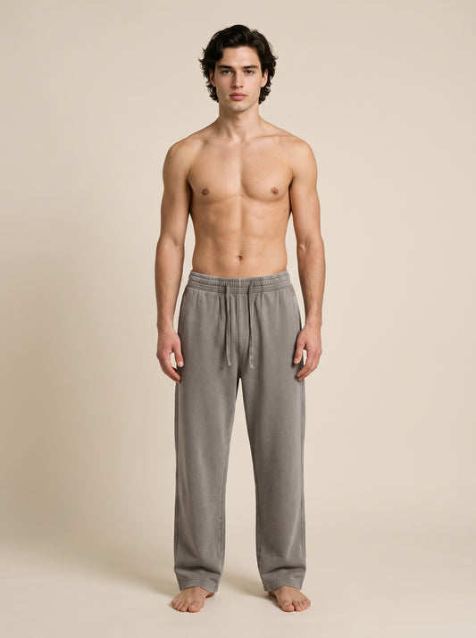 A shirtless male model stands barefoot against a plain beige studio background, wearing loose gray drawstring lounge pants.