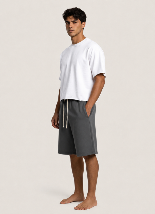 A man wearing a white crewneck t-shirt and dark gray drawstring shorts, standing barefoot against a light beige studio background.