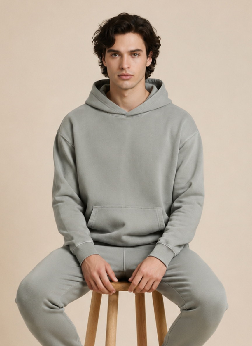 A man wearing a matching light gray hoodie and sweatpants set, seated on a wooden stool against a plain beige studio background.