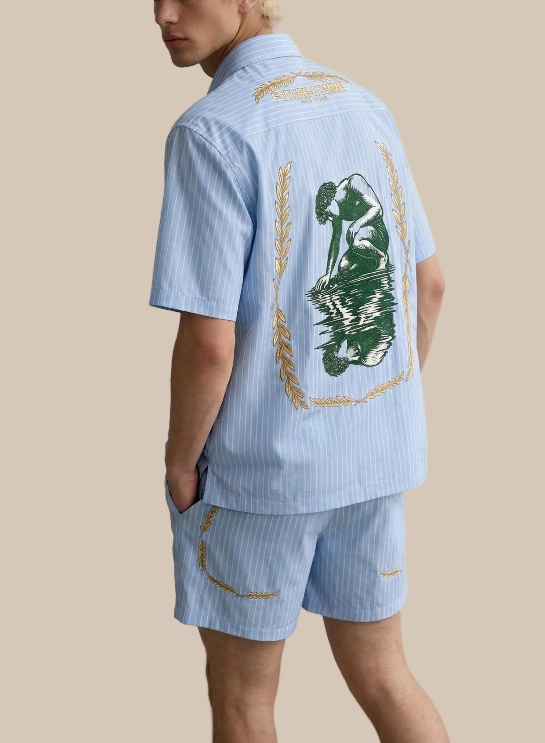 Person wearing a light blue striped shirt with a green leaf design on the back, standing against a white background.