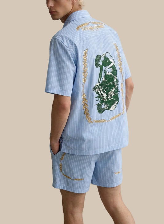 Person wearing a light blue striped shirt with a green leaf design on the back, standing against a white background.