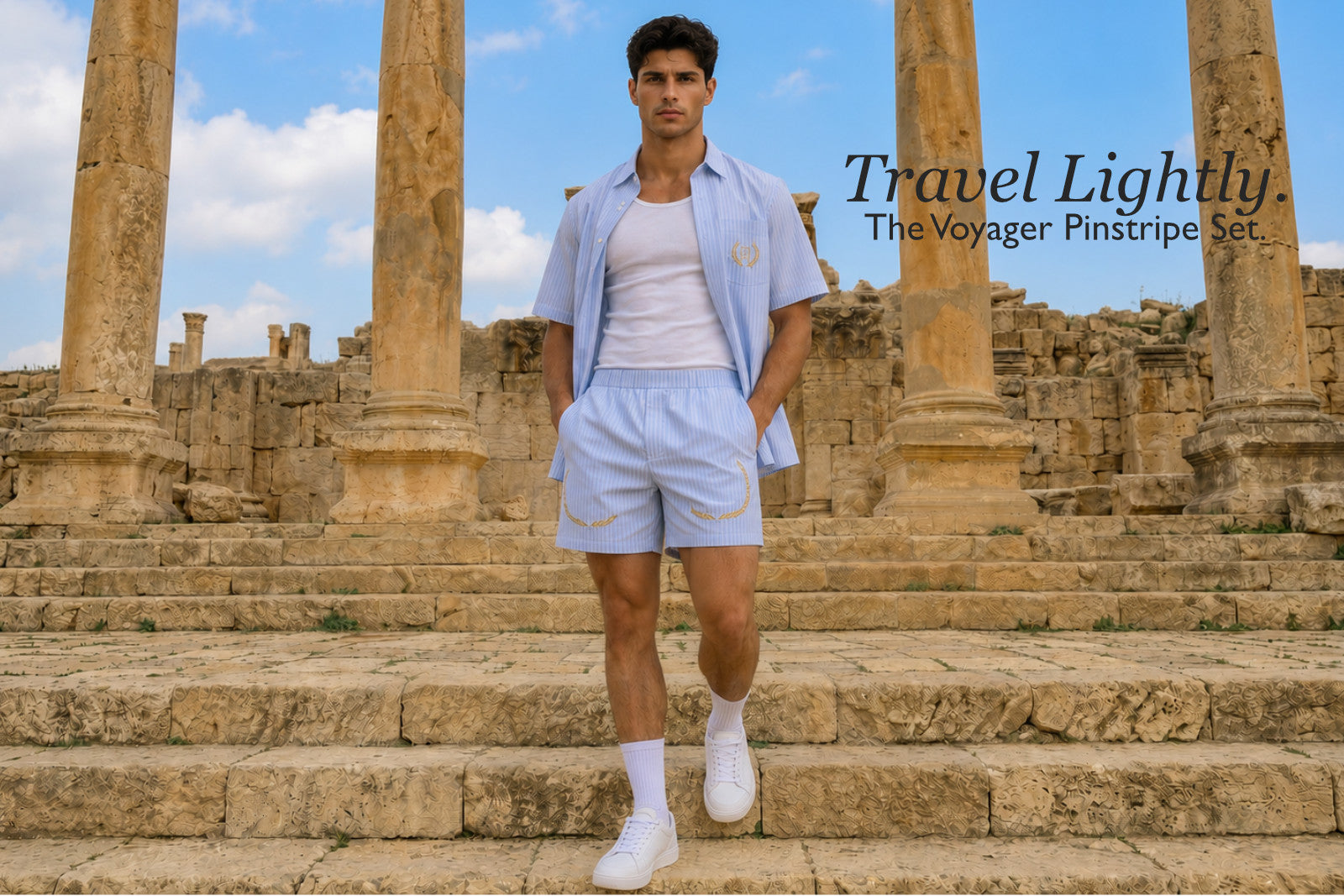 Man standing on stone steps with ancient columns in the background, wearing a light blue pinstripe shirt and shorts set. Text overlay reads, Travel Lightly. The Voyager Pinstripe Set. 