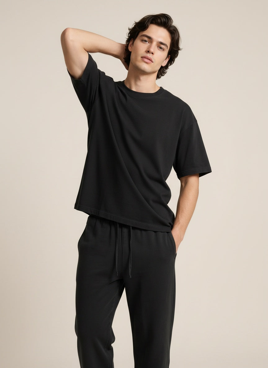 A man modeling a black short-sleeve crewneck t-shirt and matching black sweatpants against a solid light beige background.