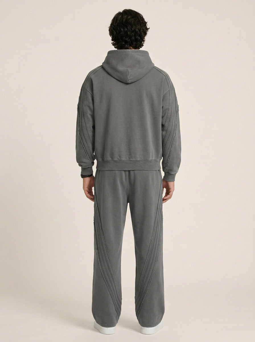 Backside of a model wearing a sun bleached deconstructed dark gray hoodie with matching sweatpants on a beige background. 