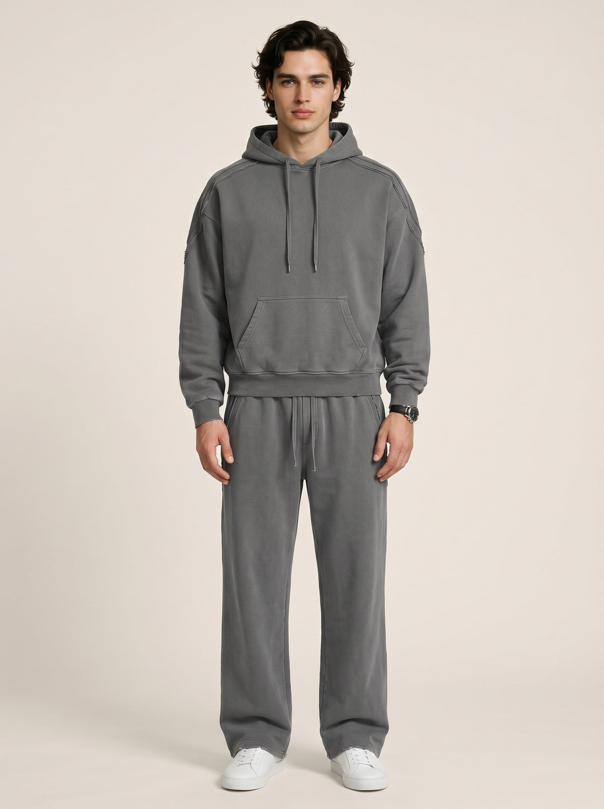 A model wearing a sun bleached deconstructed hoodie in dark gray with matching sweatpants on a beige background. 
