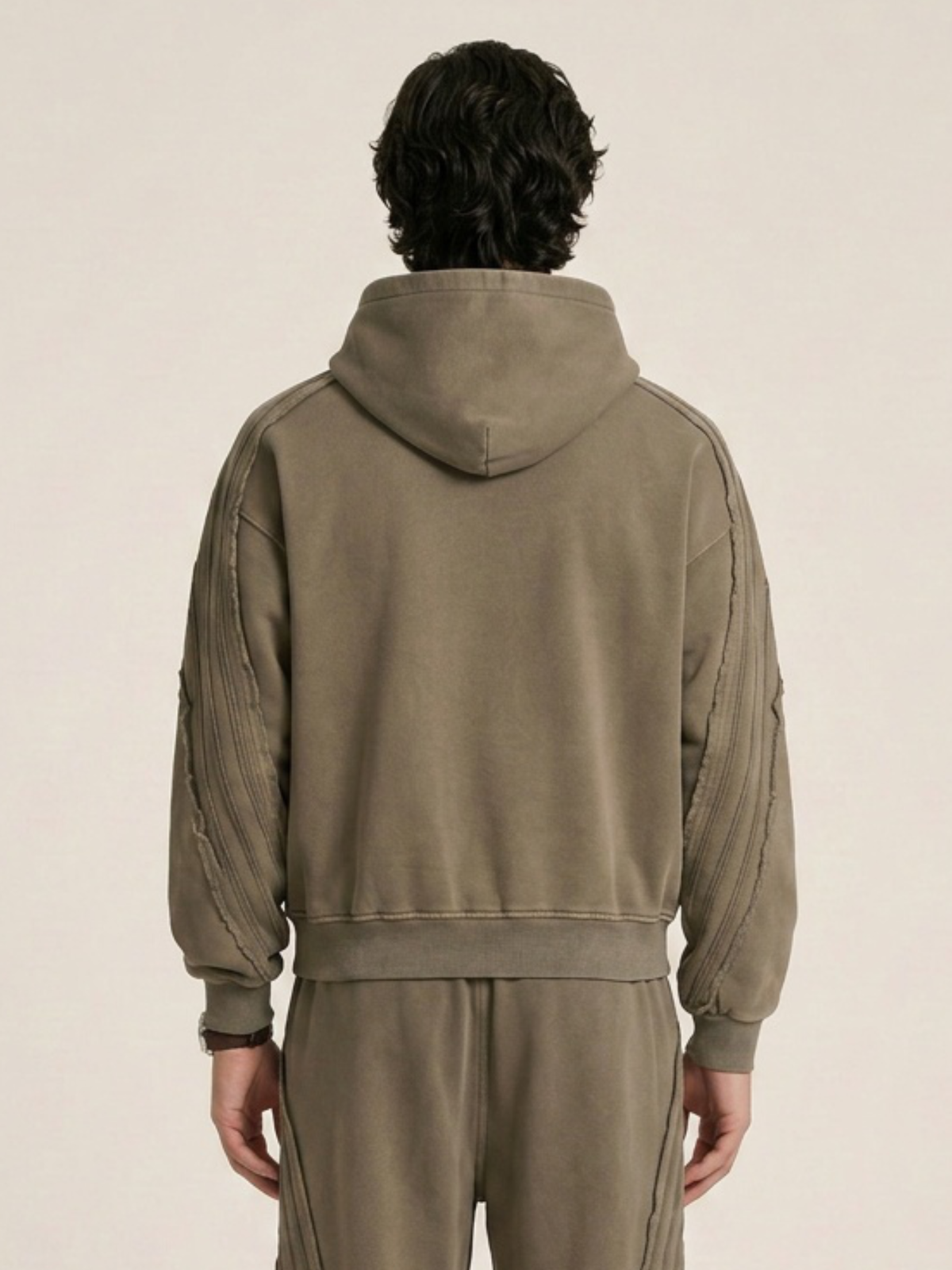 Backside of a model wearing a sun bleached deconstructed khaki hoodie with matching sweatpants on a beige background. 