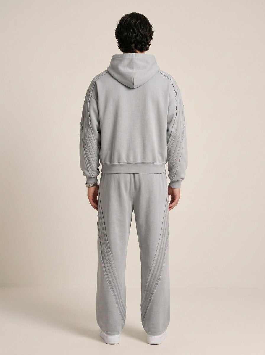 Backside of a model wearing a sun bleached deconstructed hoodie in light gray with matching sweatpants on a beige background. 