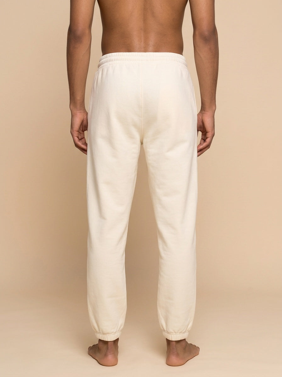 A man viewed from the back wearing cream-colored sweatpants, standing barefoot against a solid beige background.