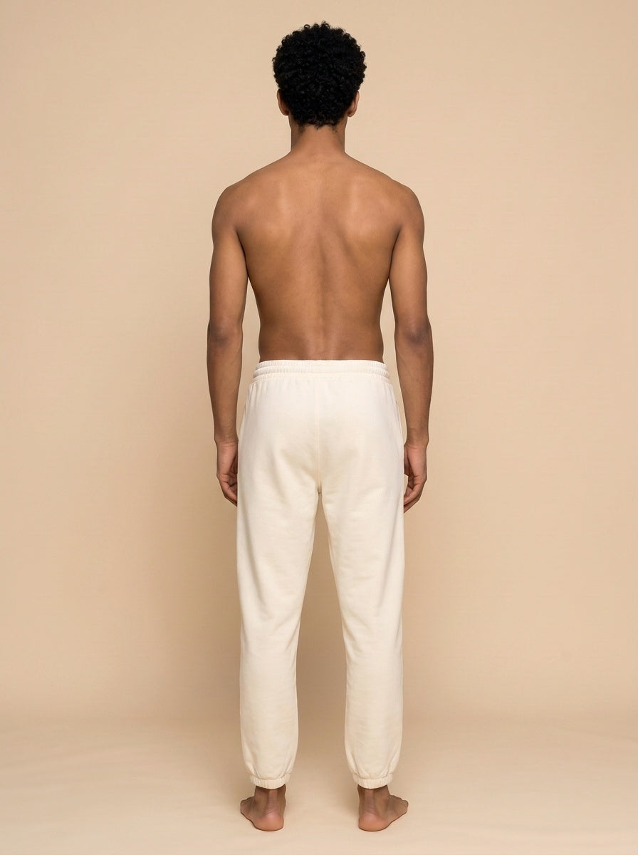 A man viewed from behind wearing loose off-white trousers, standing barefoot against a plain beige studio background.