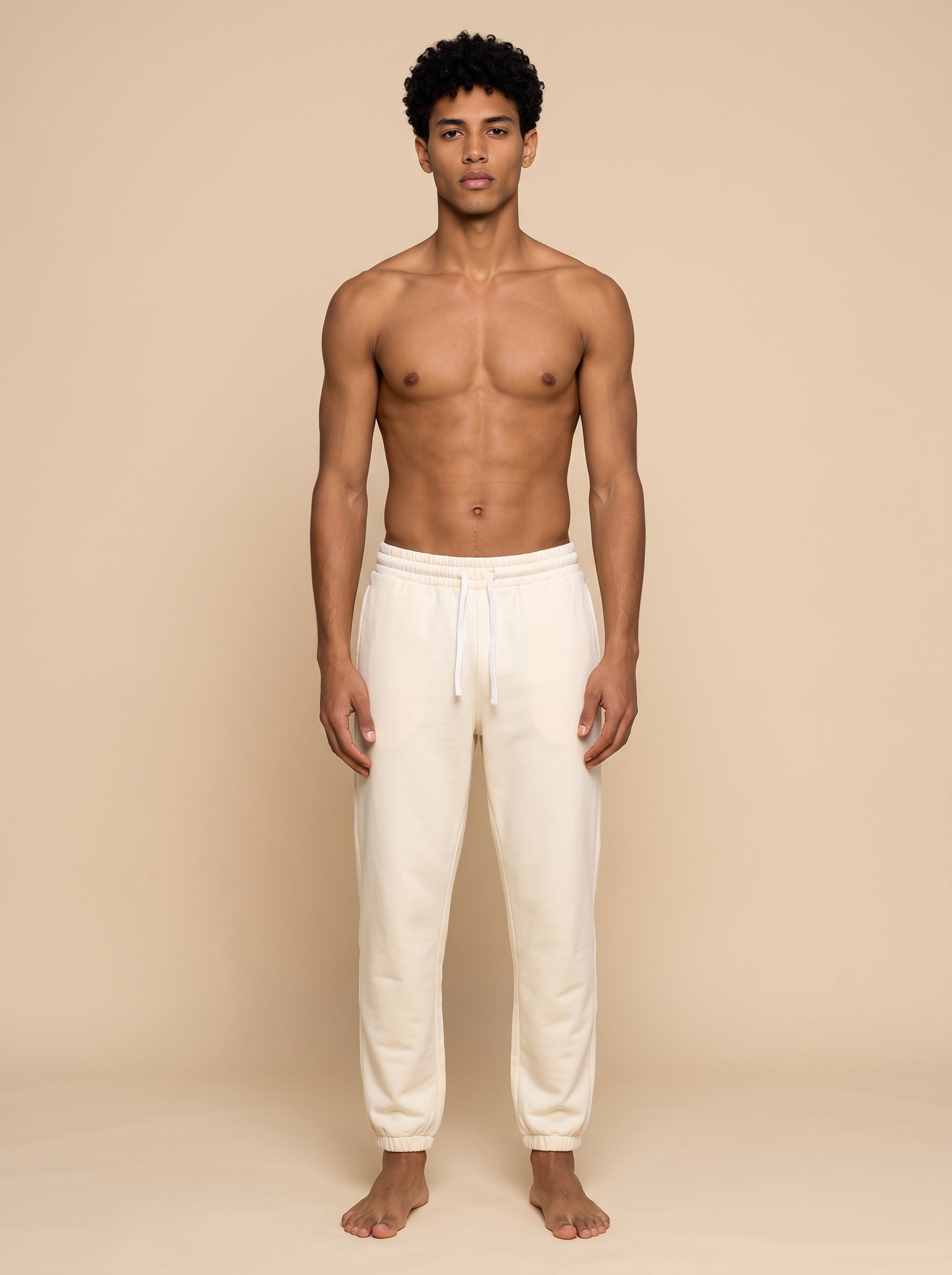 A shirtless man stands facing forward wearing cream-colored sweatpants against a plain beige studio background.