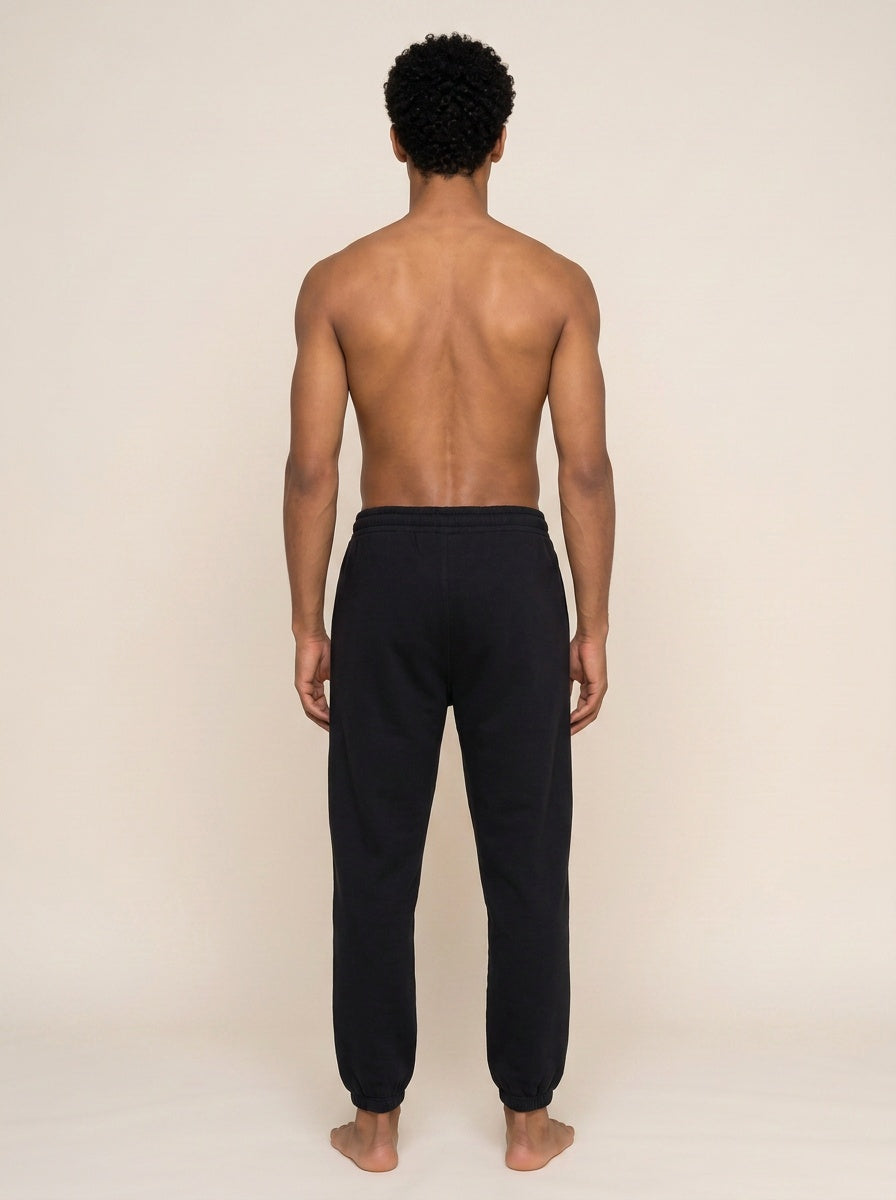 A man viewed from behind wearing black sweatpants, standing barefoot against a plain beige studio background.