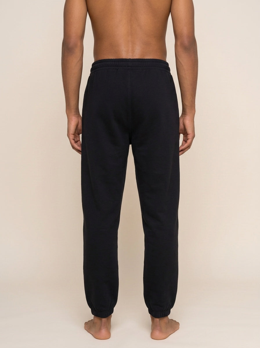 Rear view of a man wearing black sweatpants against a plain beige studio background.