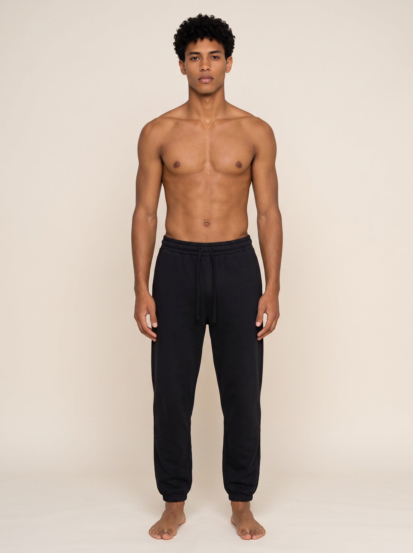 A shirtless male model wearing black sweatpants, standing against a plain beige studio background.