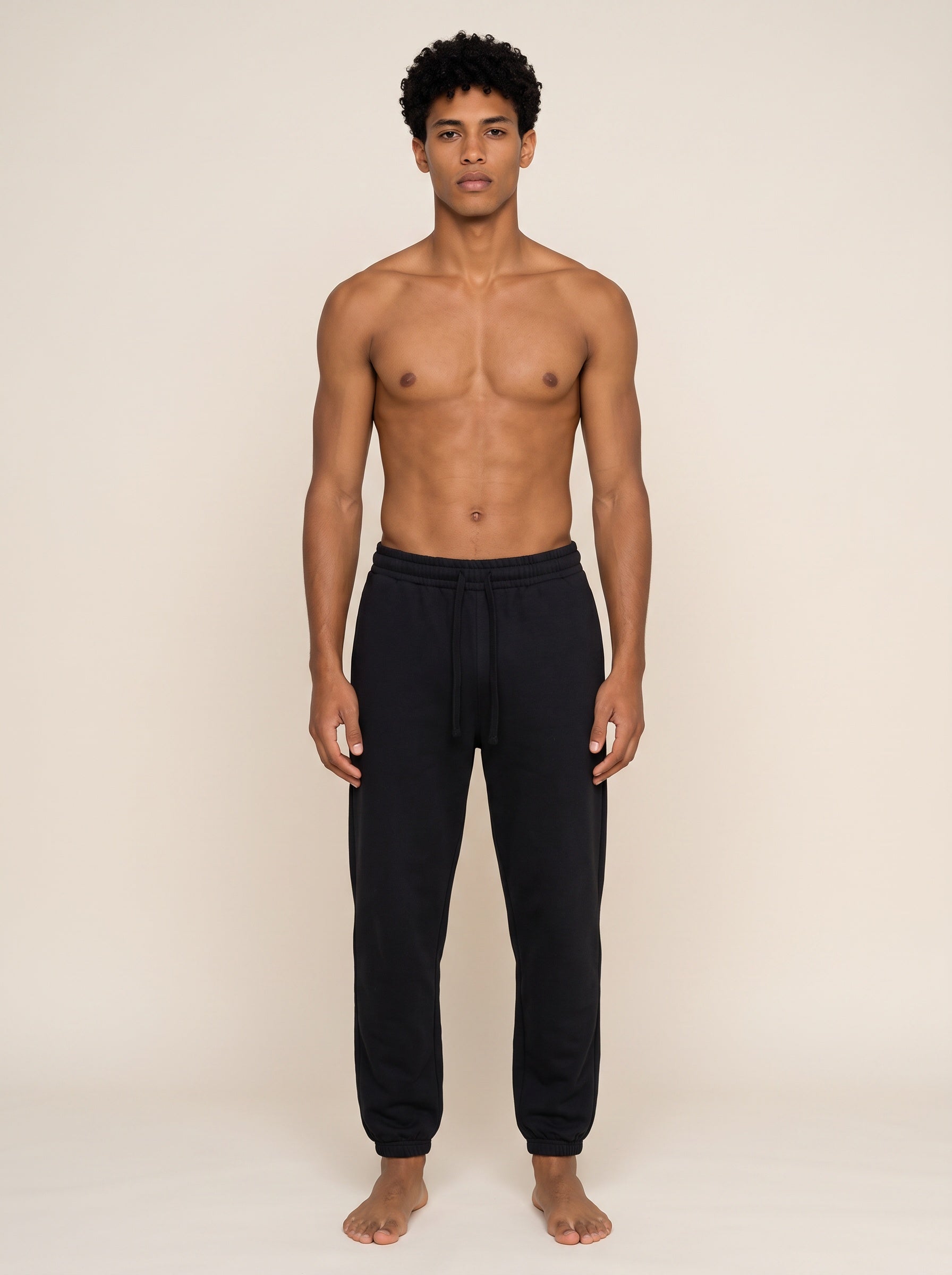 A shirtless male model wearing black sweatpants, standing against a plain beige studio background.