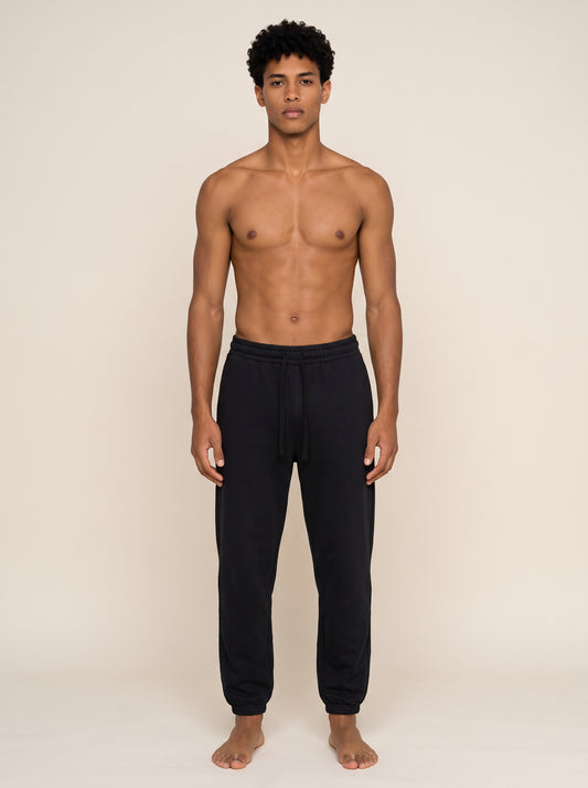 A shirtless male model wearing black sweatpants, standing against a plain beige studio background.