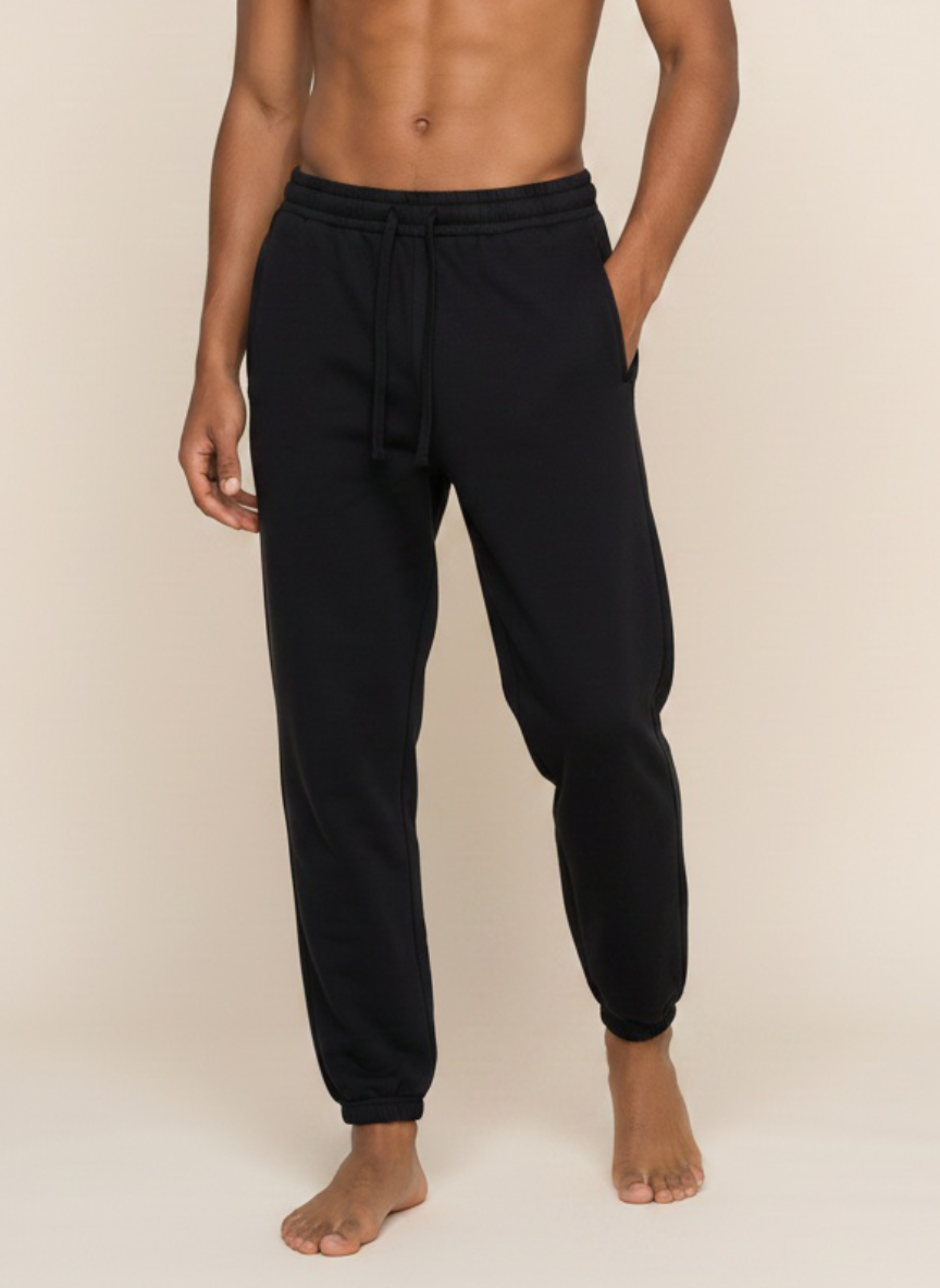 A man wearing black jogger sweatpants with a drawstring waistband, shown from the chest down against a plain beige studio background.