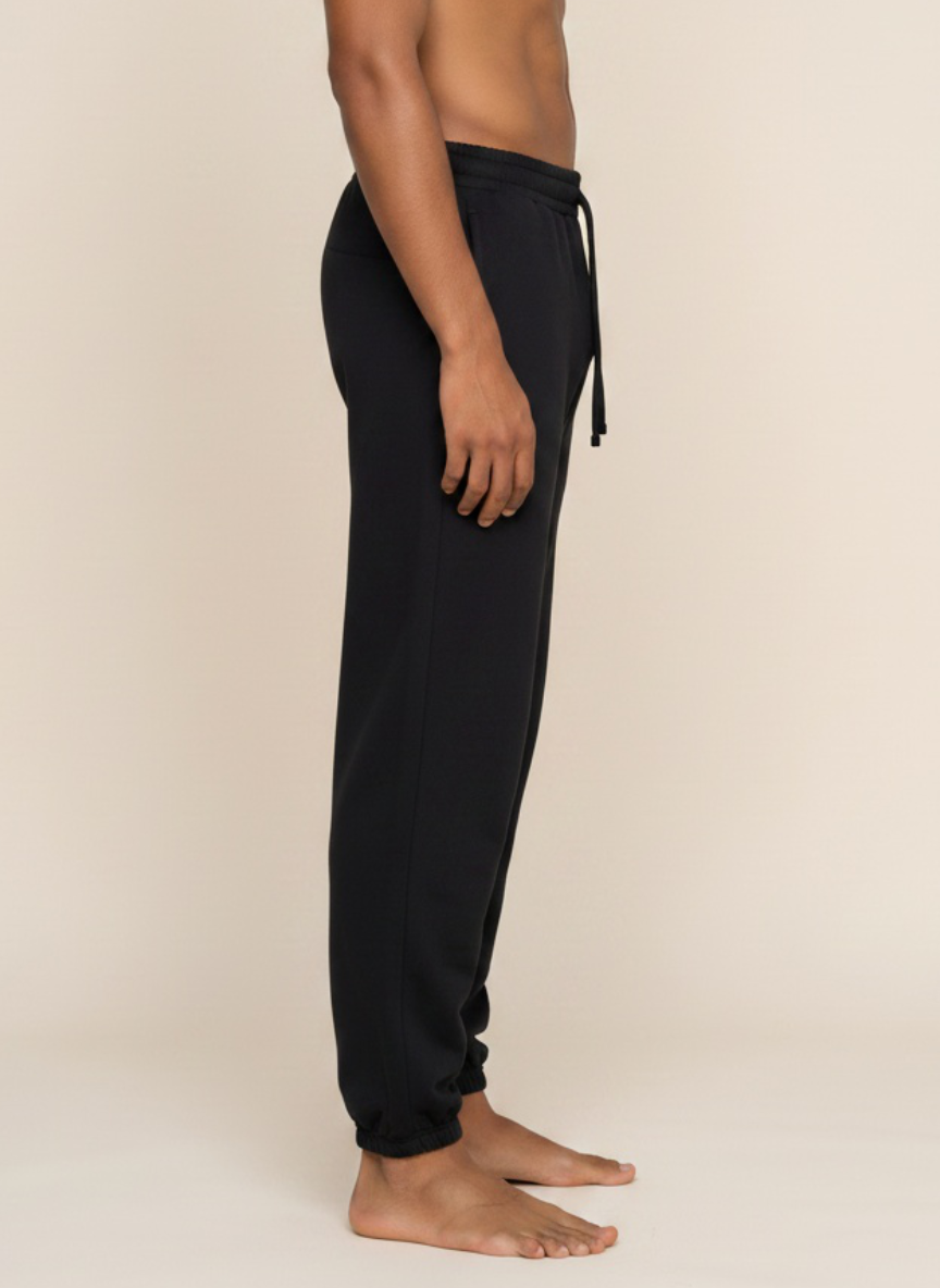 A side-profile view of a person wearing black jogger sweatpants with a drawstring waist, standing barefoot against a plain beige studio background.