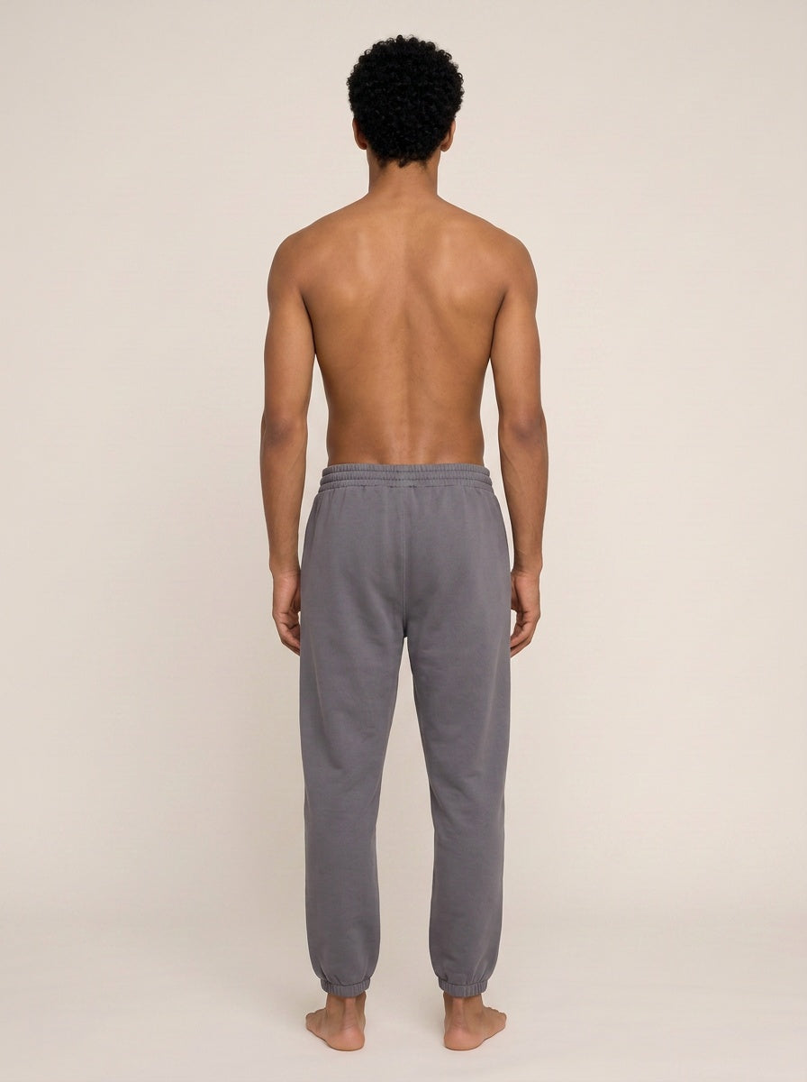 A man viewed from behind wearing gray sweatpants, standing barefoot against a plain light beige studio background.