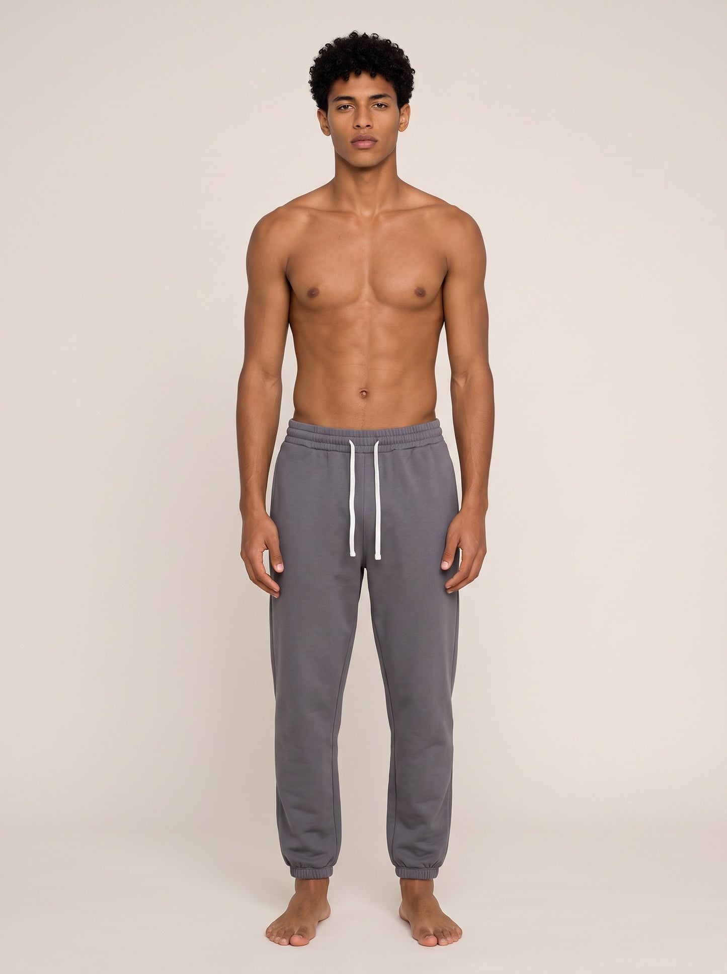 A shirtless male model stands against a plain beige studio background wearing gray jogger sweatpants with a white drawstring.