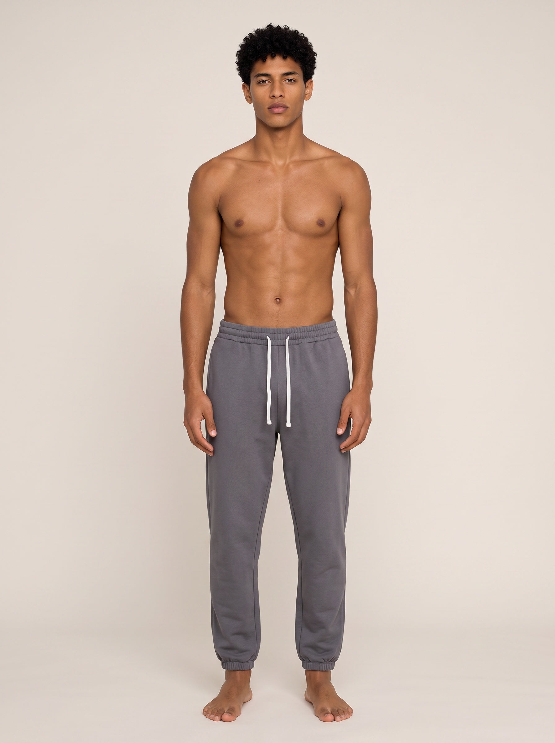 A shirtless male model stands against a plain beige studio background wearing gray jogger sweatpants with a white drawstring.