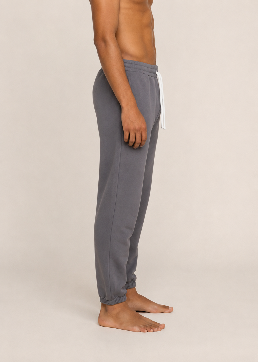 A side-profile view of a person wearing gray jogger sweatpants with a white drawstring, standing barefoot against a plain light beige studio background.