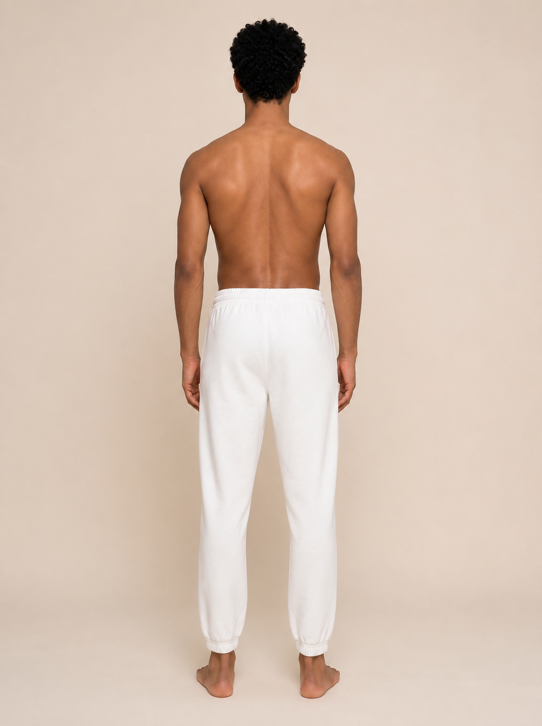 A man viewed from behind wearing white jogger-style pants, standing barefoot against a plain beige studio background.