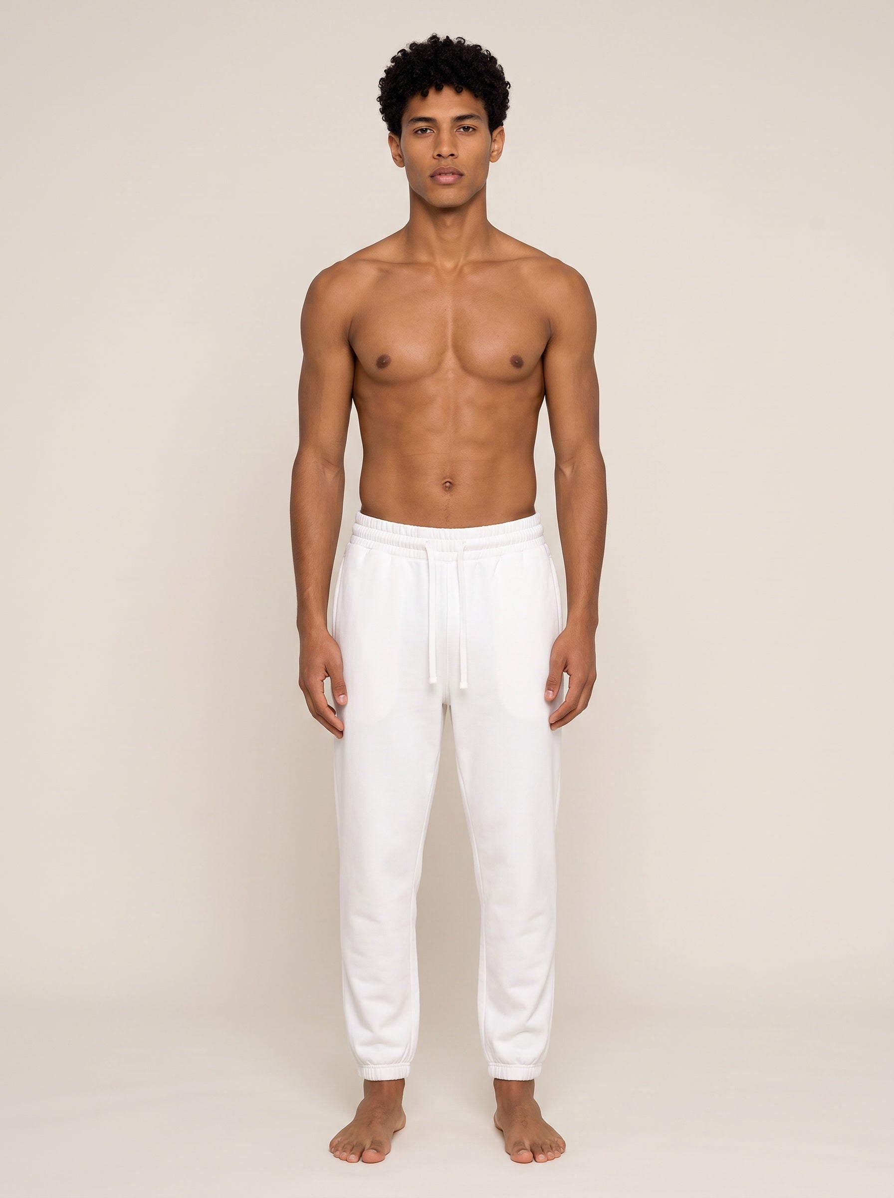 A shirtless male model wearing white drawstring sweatpants, standing against a plain beige studio background.