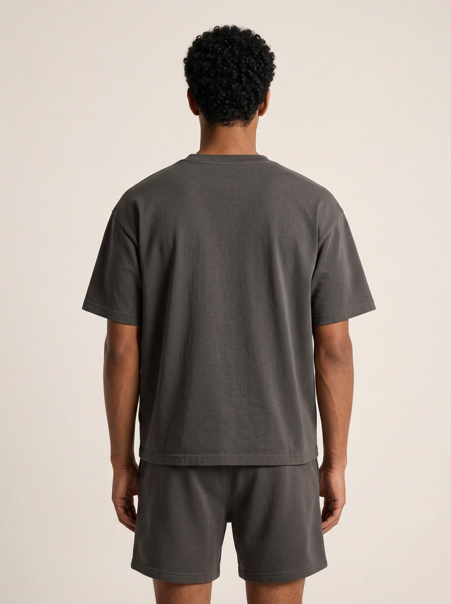 A man viewed from behind wearing a dark gray short-sleeve t-shirt and matching dark gray shorts against a plain beige studio background.