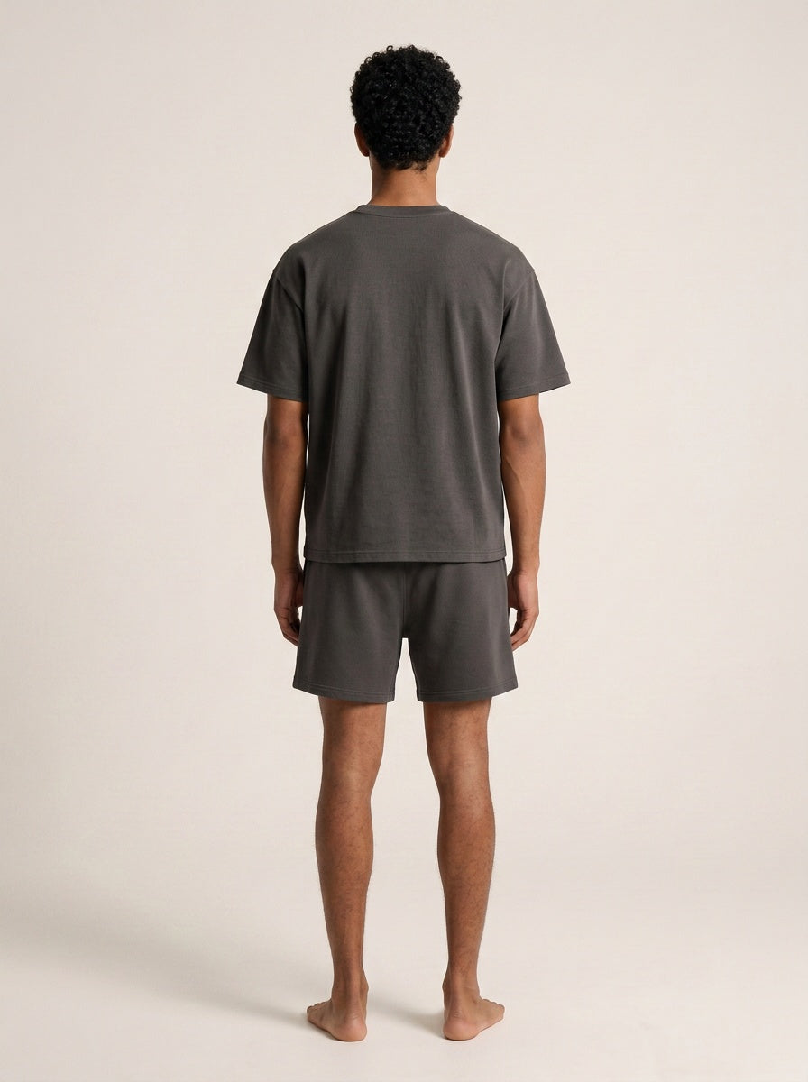 A man viewed from behind wearing a dark gray short-sleeve t-shirt and matching dark gray shorts against a plain beige studio background.