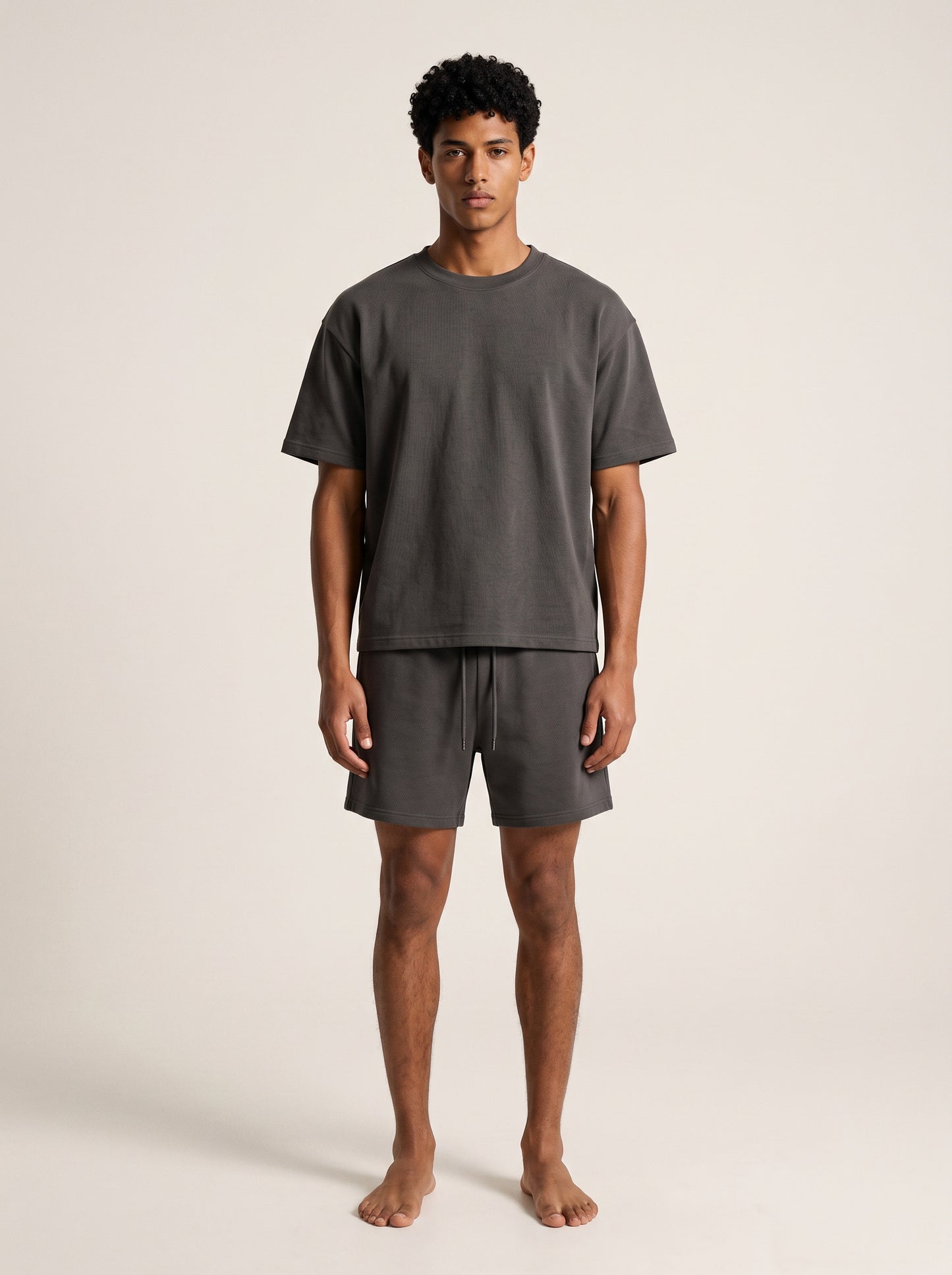 A man modeling a dark gray short-sleeve t-shirt and matching dark gray shorts against a plain, light beige studio background.