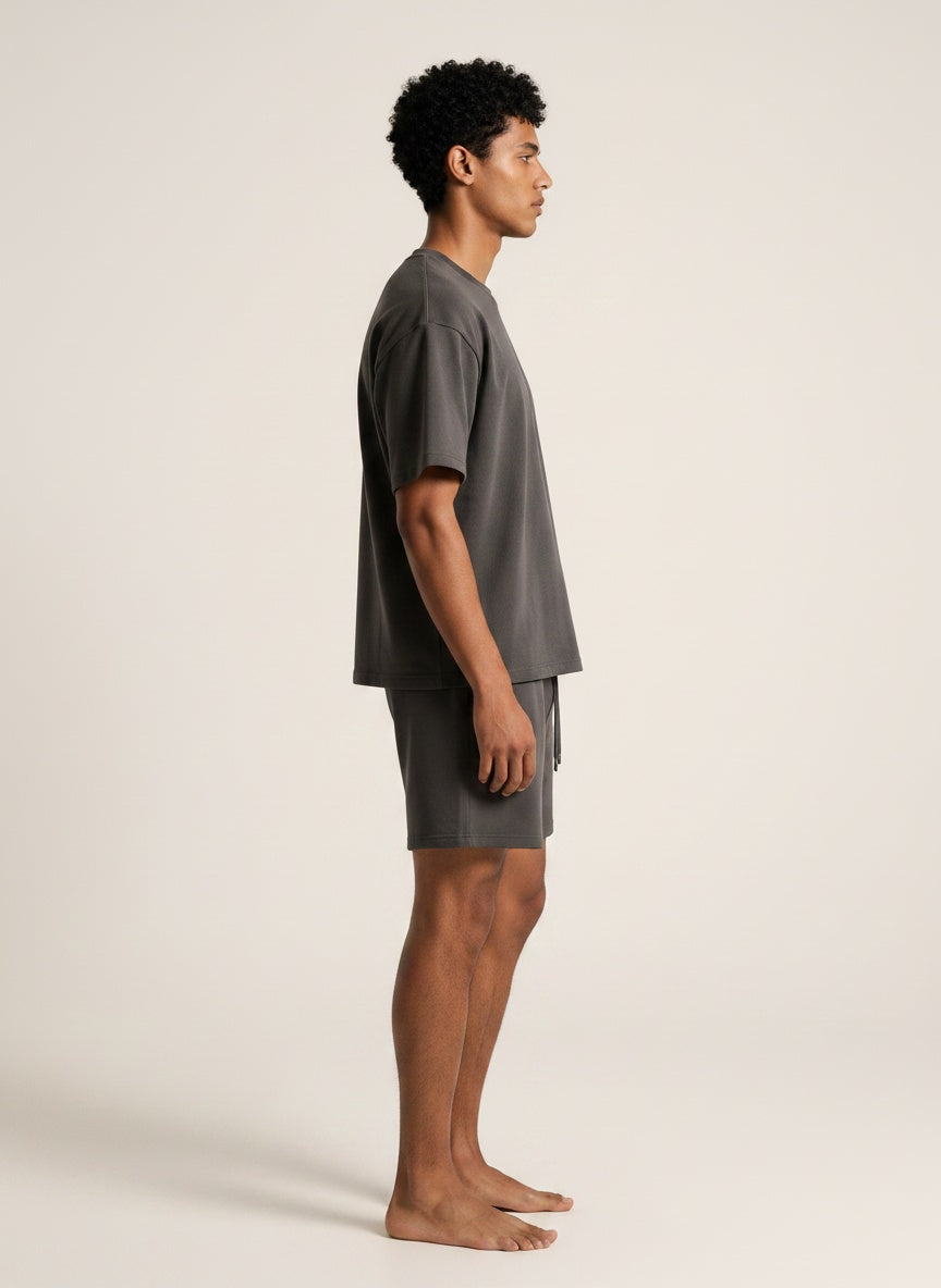 A man in profile wearing a dark gray oversized t-shirt and matching drawstring shorts, standing barefoot against a plain light beige studio background.