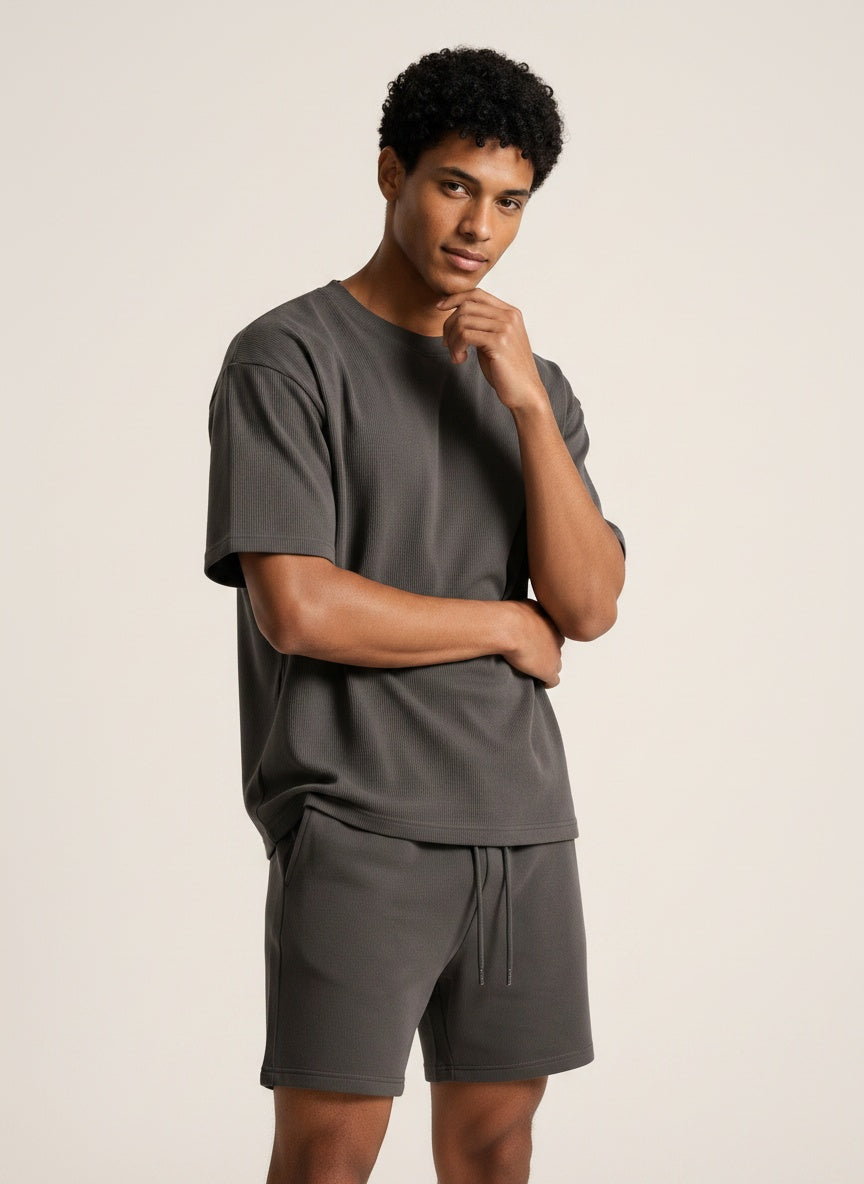 A man modeling a dark gray short-sleeve t-shirt and matching drawstring shorts against a plain beige studio background.
