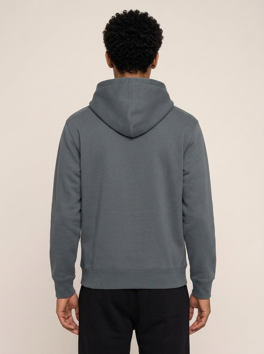 A man viewed from behind wearing a dark gray pullover hoodie and black pants, standing against a plain light beige studio background.
