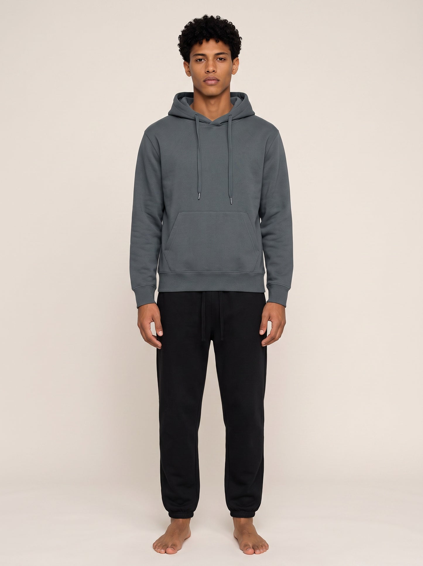 A man wearing a dark gray pullover hoodie and black sweatpants, standing barefoot against a plain beige studio background.