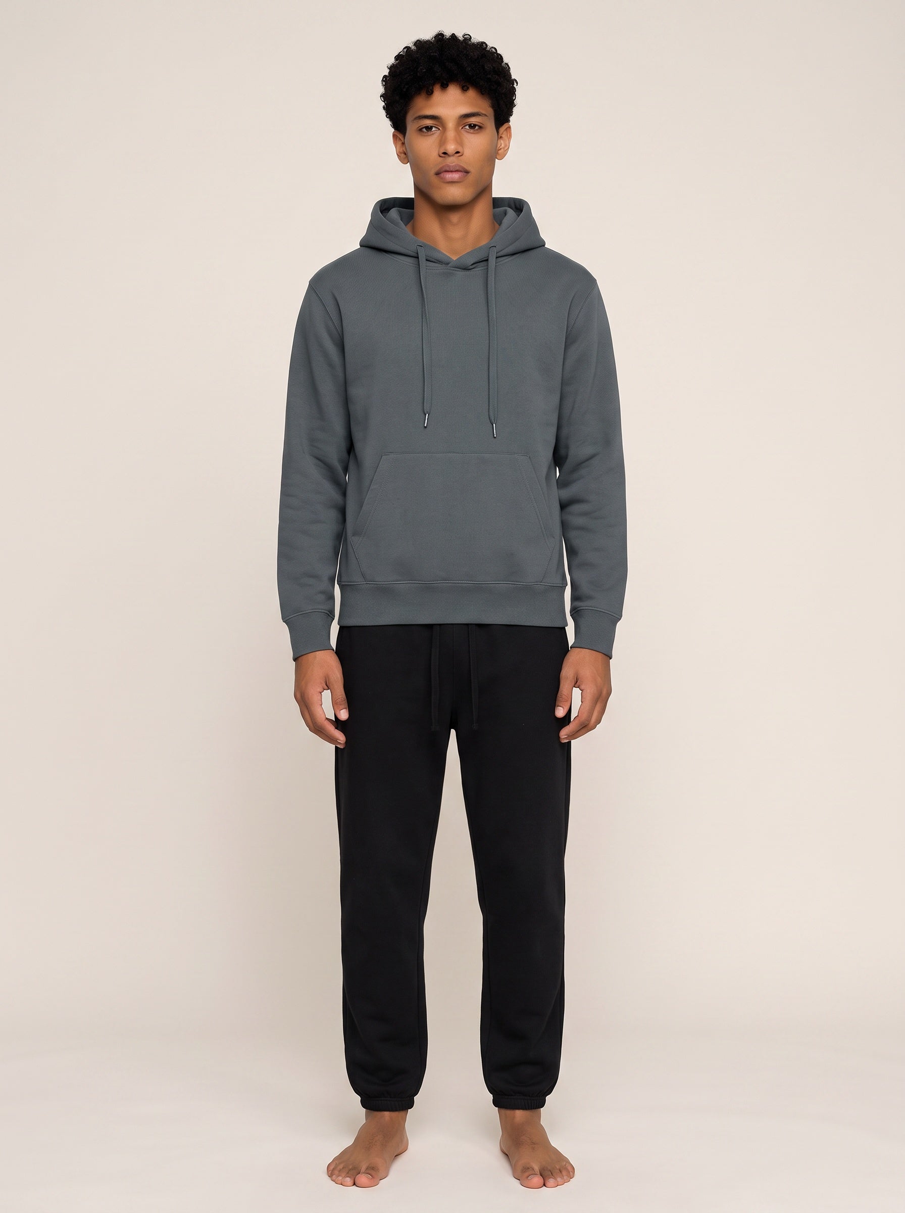 A man wearing a dark gray pullover hoodie and black sweatpants, standing barefoot against a plain beige studio background.