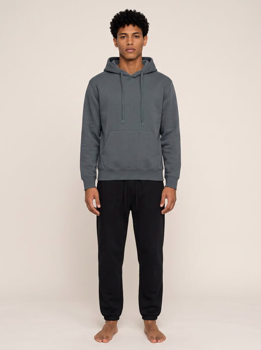 A man wearing a dark gray pullover hoodie and black sweatpants, standing barefoot against a plain beige studio background.
