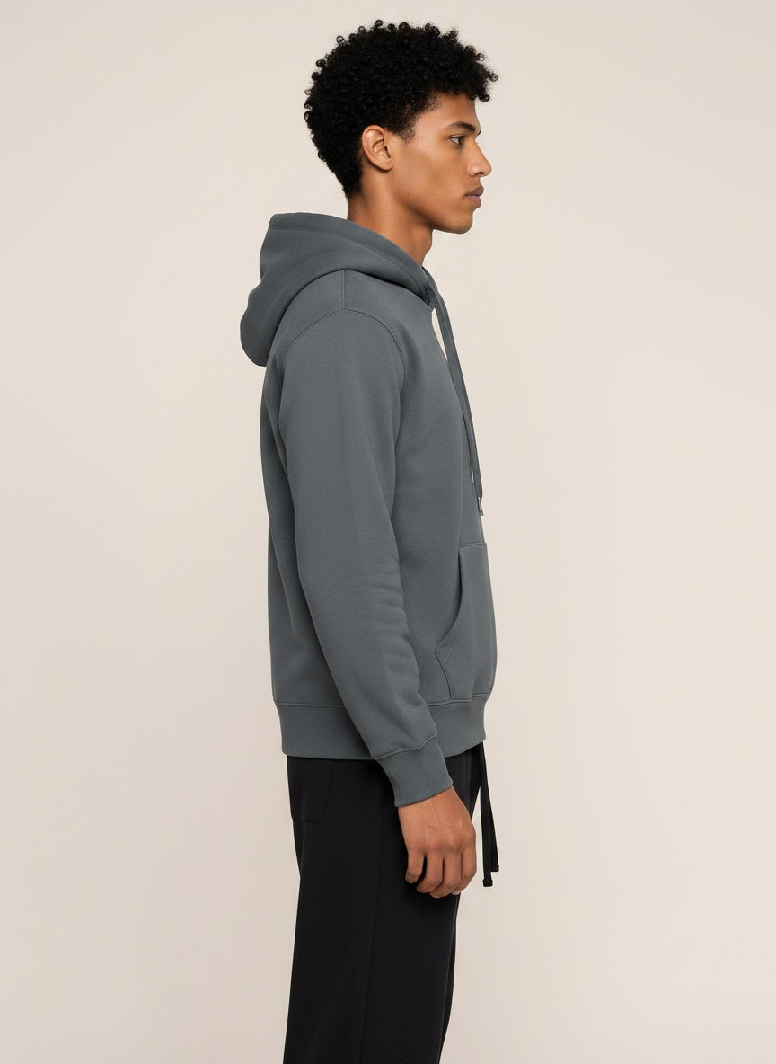 A man in profile wearing a dark gray pullover hoodie and black pants, standing against a plain light beige studio background.