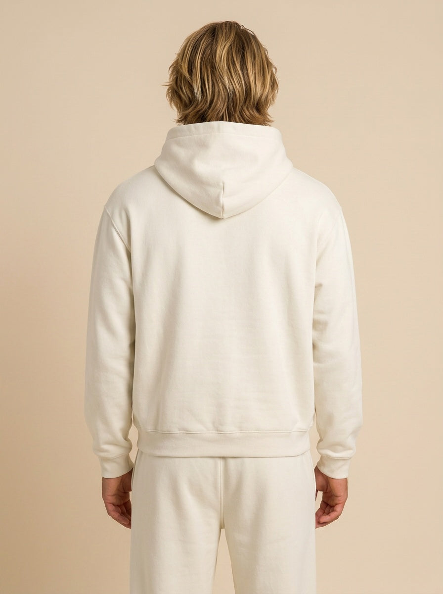 A person shown from the back wearing a plain hooded sweatshirt and matching sweatpants in a warm off-white or cream color, against a solid beige studio background.