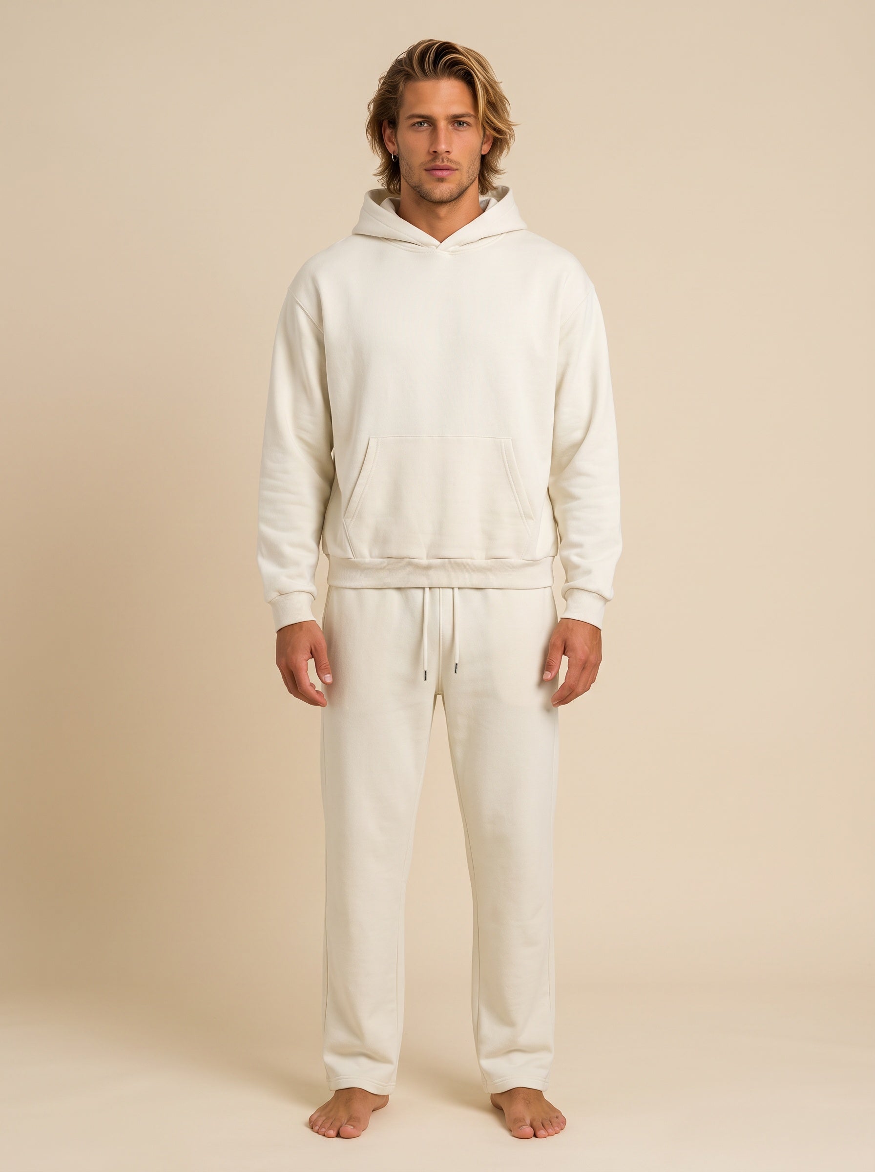 A man modeling a matching off-white hooded sweatshirt and sweatpants lounge set against a solid beige studio background.