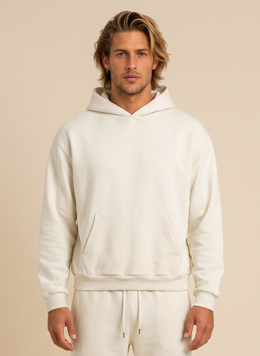 A man wearing a cream-colored pullover hoodie and matching sweatpants, standing against a solid beige background.