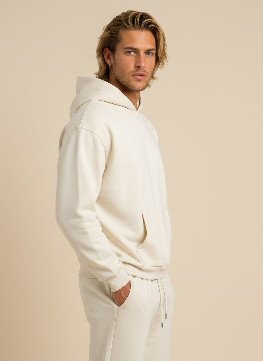 A man wearing a matching off-white fleece hoodie and jogger sweatpants set, shown in profile against a solid beige studio background.