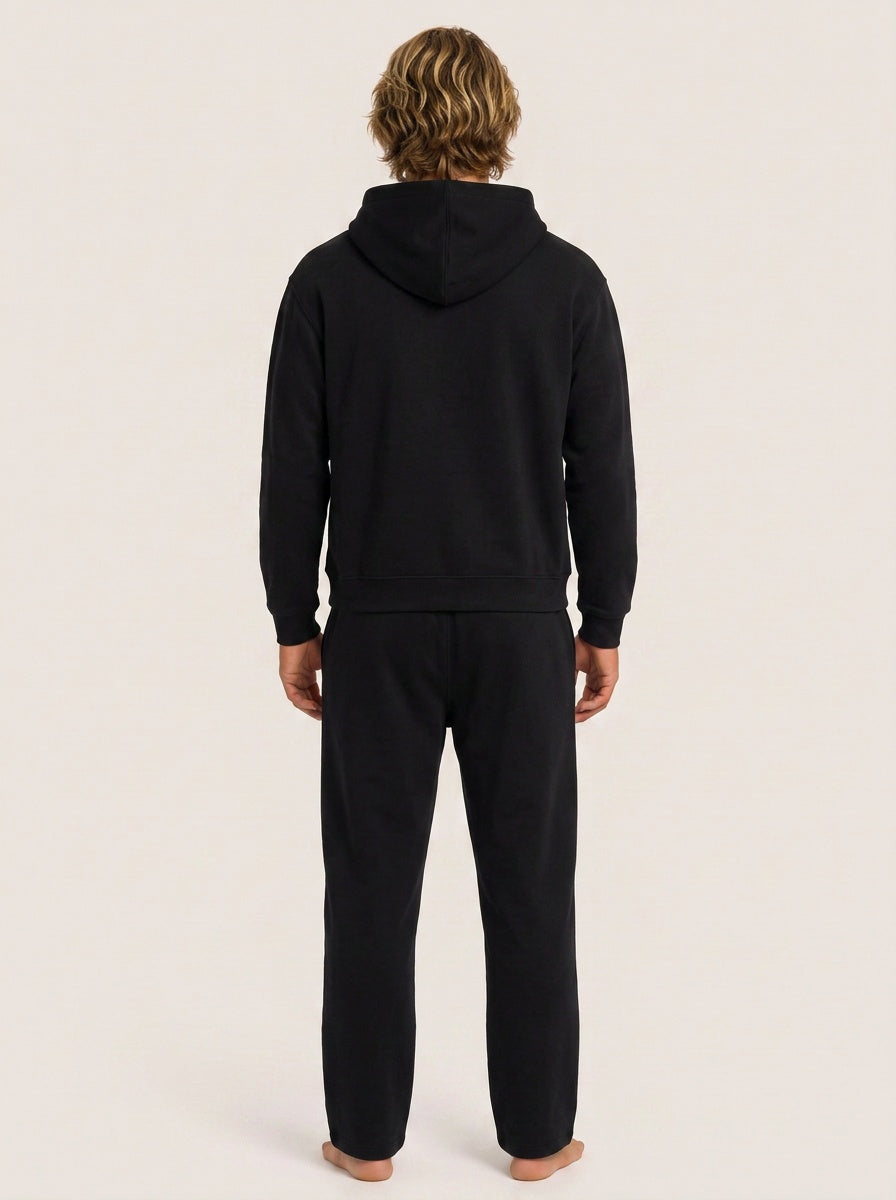 A man viewed from behind wearing a black hooded sweatshirt and matching black sweatpants against a plain light beige studio background.