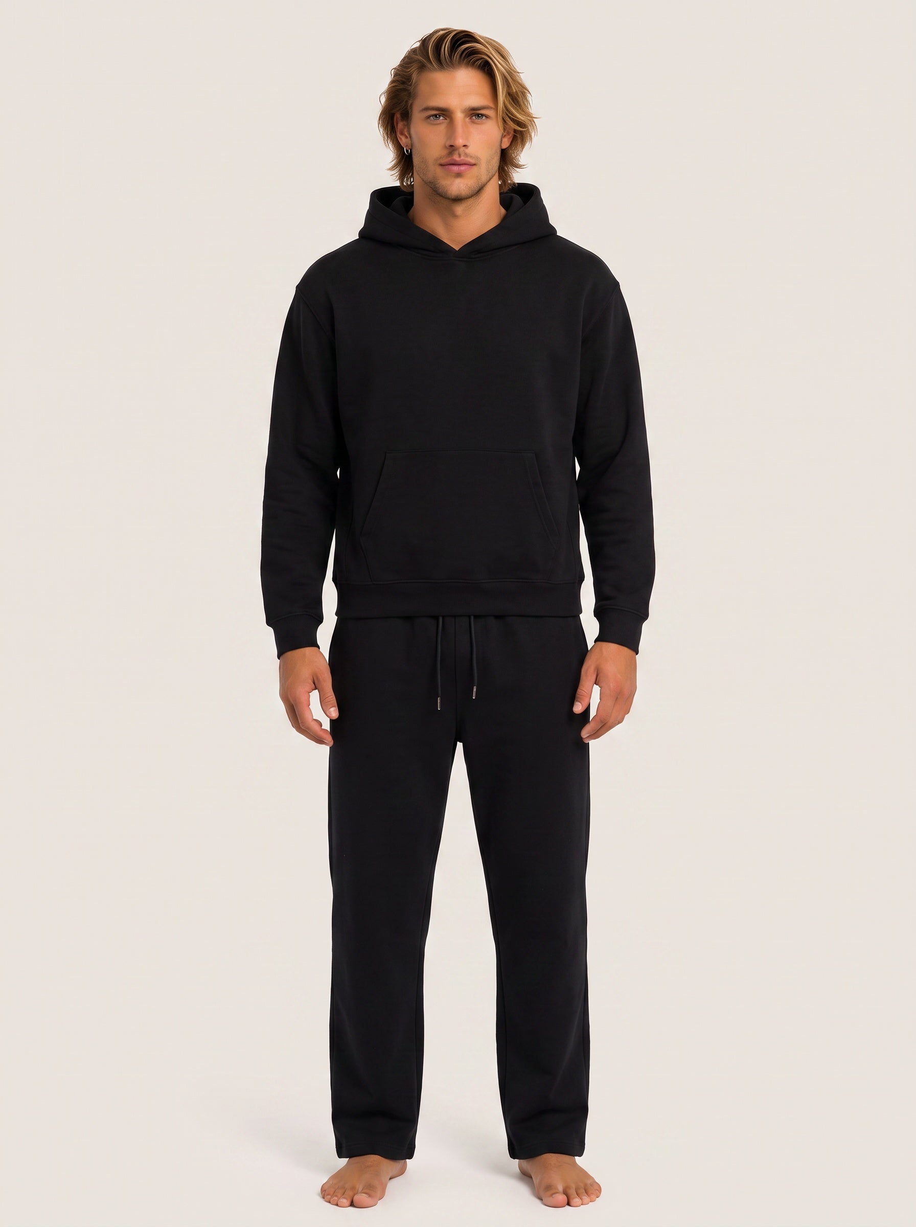 A man wearing a black pullover hoodie and matching black sweatpants, standing barefoot against a plain light beige studio background.