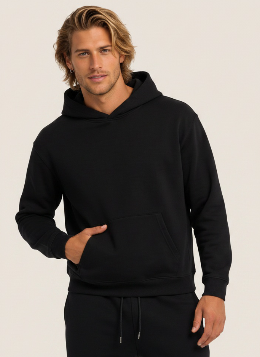 A man wearing a black pullover hoodie with a front kangaroo pocket and matching black sweatpants, standing against a plain light beige studio background.
