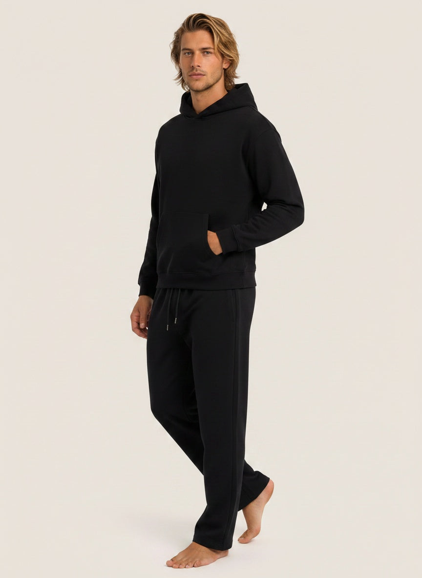 A man modeling a black two-piece loungewear set consisting of a long-sleeve pullover hoodie and matching sweatpants, standing against a solid light beige background.