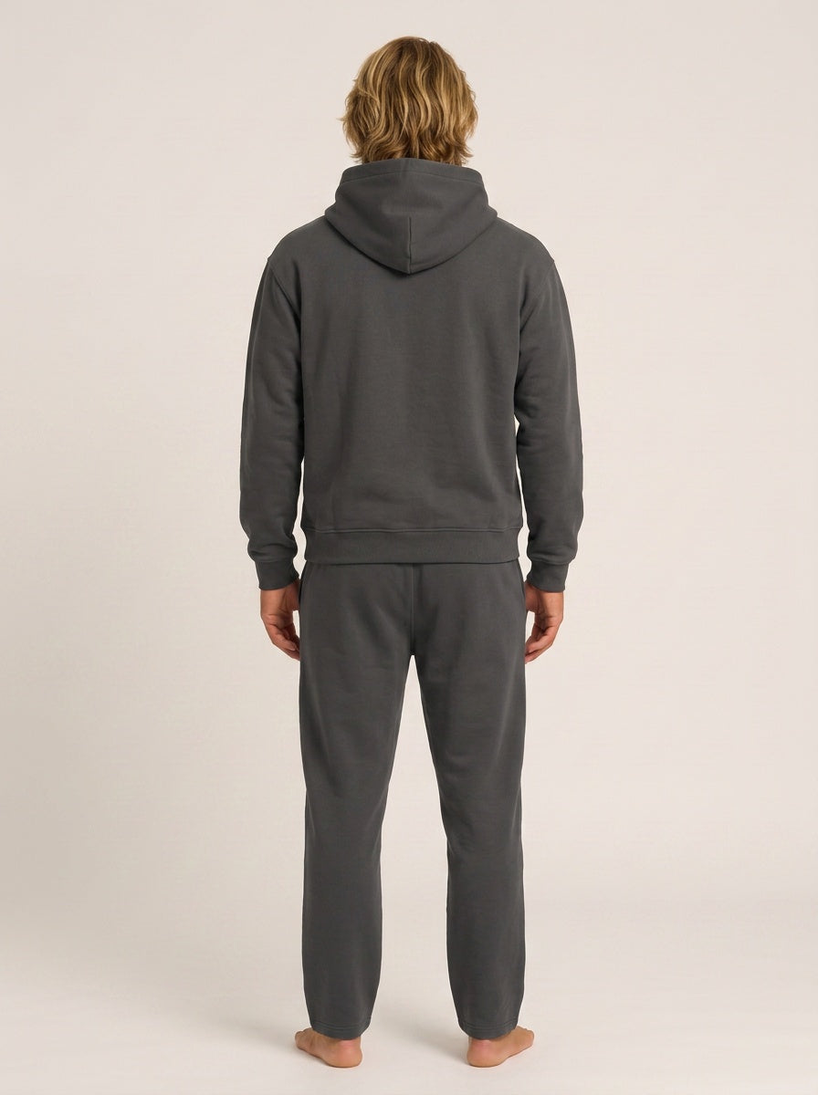 A man viewed from behind wearing a dark gray hooded sweatshirt and matching dark gray sweatpants, standing barefoot against a plain light beige studio background.