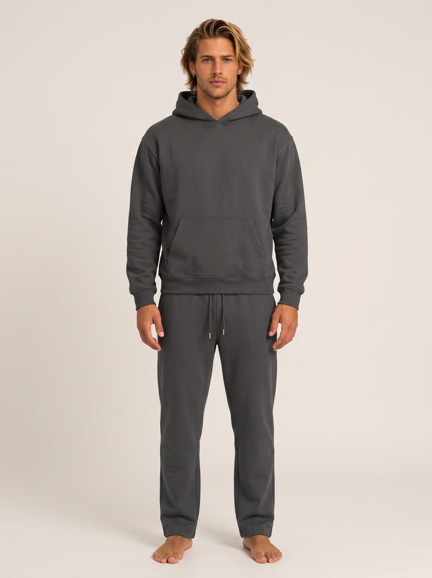 A man modeling a dark gray two-piece loungewear set consisting of a pullover hoodie and matching sweatpants, standing against a plain light beige studio background.