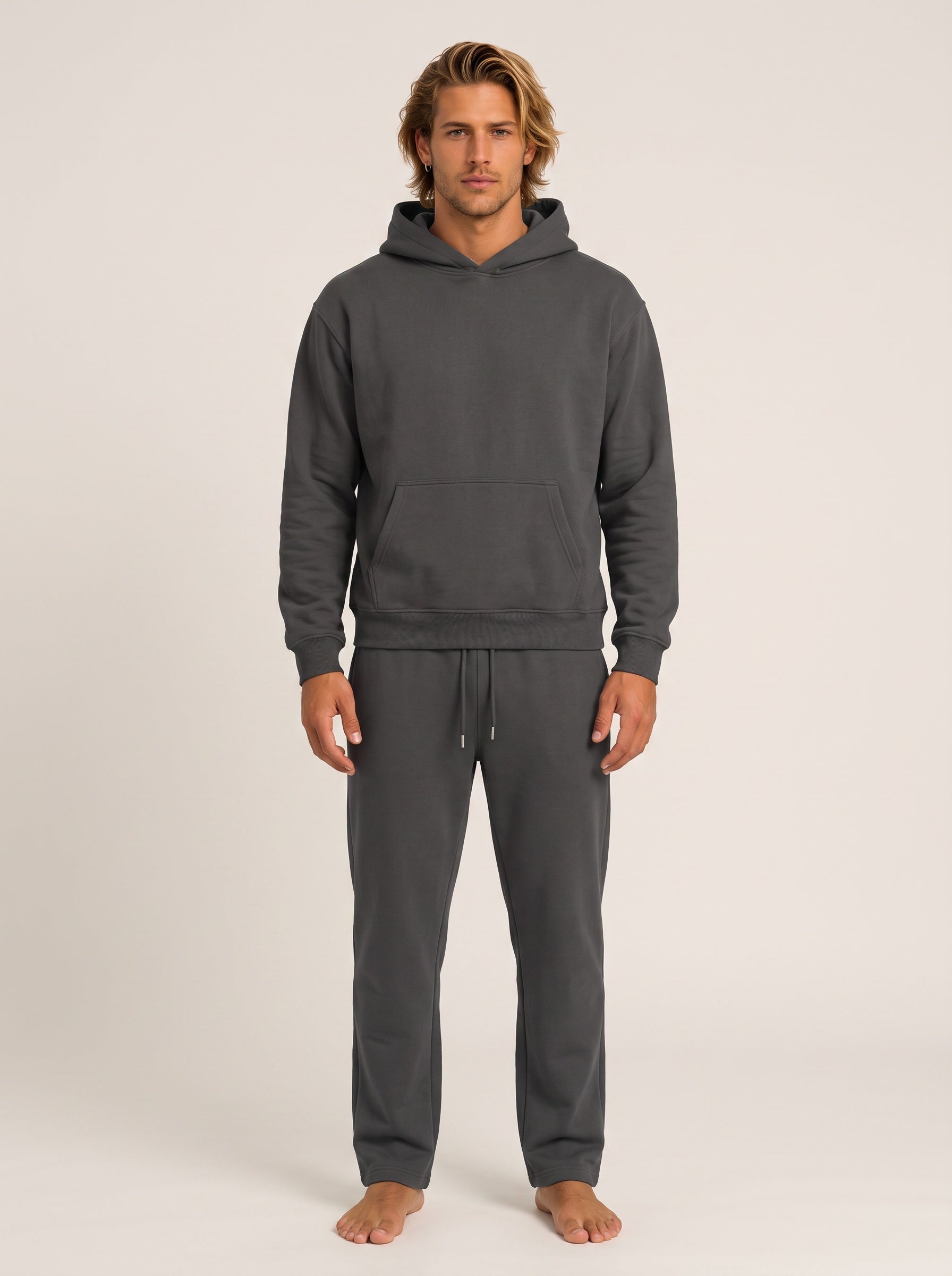 A man modeling a dark gray two-piece loungewear set consisting of a pullover hoodie and matching sweatpants, standing against a plain light beige studio background.